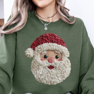 May include: Olive green sweatshirt featuring a detailed Santa Claus face design. The Santa face has a red hat, a white beard, and rosy cheeks. The sweatshirt is a solid color, perfect for the holiday season.
