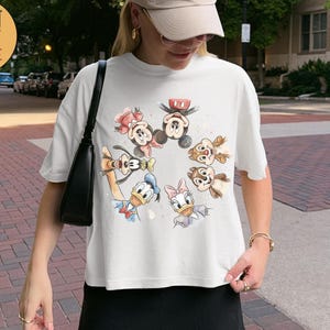 May include: White Comfort Colors Boxy Tee featuring a watercolor design of Disney characters including Mickey Mouse, Minnie Mouse, Donald Duck, Daisy Duck, Chip, and Dale. The shirt has short sleeves and a relaxed fit.
