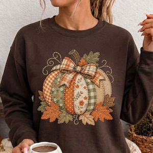 May include: A dark chocolate brown long-sleeve sweatshirt featuring a central graphic of a patchwork-style pumpkin. The pumpkin is composed of various fabric patterns including plaid, gingham, and floral prints, topped with a plaid bow and autumn leaves.
