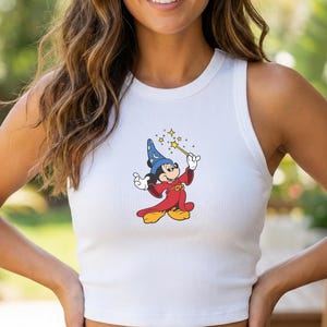 May include: White ribbed tank top featuring a colorful cartoon graphic of Mickey Mouse dressed as a wizard, holding a wand with stars. The sleeveless top is paired with blue denim jeans.