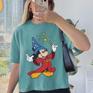 K&ouml;nnte beinhalten: T&uuml;rkisfarbenes Comfort Colors Boxy-T-Shirt mit einer Grafik von Mickey Mouse als Zauberer, der einen Zauberstab mit Sternen h&auml;lt. Die Zeichentrickfigur tr&auml;gt einen blauen Zaubererhut und eine rote Robe. Auf dem Shirt steht "Chalky Mint".
