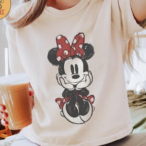 May include: Ivory-colored Comfort Colors Boxy Tee featuring a distressed Minnie Mouse graphic. The graphic shows Minnie Mouse with a red polka dot bow and matching accents. The shirt is a relaxed fit.
