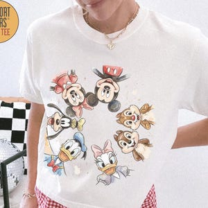 May include: Ivory-colored Comfort Colors boxy tee featuring a watercolor design of Mickey Mouse, Minnie Mouse, Donald Duck, Daisy Duck, Goofy, and Chip and Dale. The shirt has a crew neck and short sleeves. The shirt is paired with red and white checkered shorts.