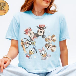 May include: Light blue Comfort Colors boxy tee featuring a watercolor design of Disney characters including Mickey Mouse, Minnie Mouse, Donald Duck, Daisy Duck, Goofy, and Chip and Dale. The shirt has short sleeves.