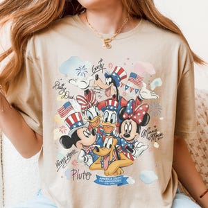 May include: Tan t-shirt featuring a colorful graphic of Disney characters Mickey Mouse, Minnie Mouse, Donald Duck, Daisy Duck, Goofy, and Pluto, celebrating the USA's 250th anniversary. The design includes American flags and fireworks.