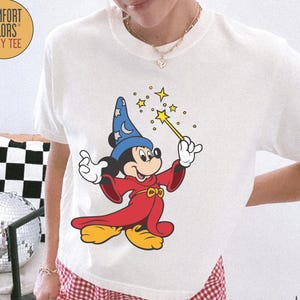 K&ouml;nnte beinhalten: Elfenbeinfarbenes Comfort Colors Boxy Tee mit einem Mickey Mouse-Zauberer-Motiv, das einen Zauberstab mit Sternen h&auml;lt. Das T-Shirt hat einen Rundhalsausschnitt und kurze &Auml;rmel. Dazu werden rot-wei&szlig; karierte Shorts getragen.