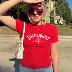 May include: A red t-shirt featuring the word "Disneyland" in white script, with "Est. 1955" and a Mickey Mouse graphic. The top is short-sleeved.