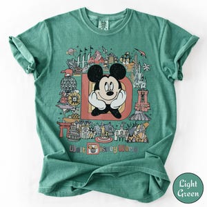 Puede incluir: Camiseta verde claro con un gr&aacute;fico colorido de Mickey Mouse, la letra "D" y varios monumentos de Disney World. El texto "Walt Disney World" est&aacute; impreso en la parte inferior del dise&ntilde;o.