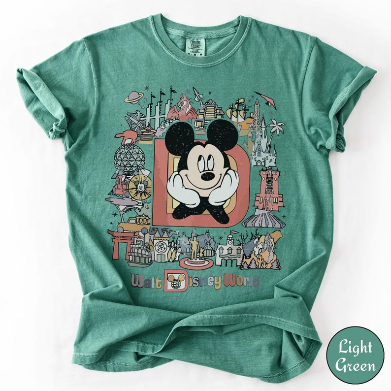 Puede incluir: Camiseta verde claro con un gr&aacute;fico colorido de Mickey Mouse, la letra "D" y varios monumentos de Disney World. El texto "Walt Disney World" est&aacute; impreso en la parte inferior del dise&ntilde;o.