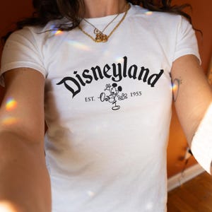 May include: White t-shirt with the word "Disneyland" in a decorative font, accompanied by "EST. 1955" and a Mickey Mouse illustration. The t-shirt has a round neck.
