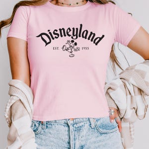 May include: A pale pink t-shirt with the word "Disneyland" in black script. Below the word is a black graphic of Mickey Mouse and the text "EST. 1955". The shirt has short sleeves.