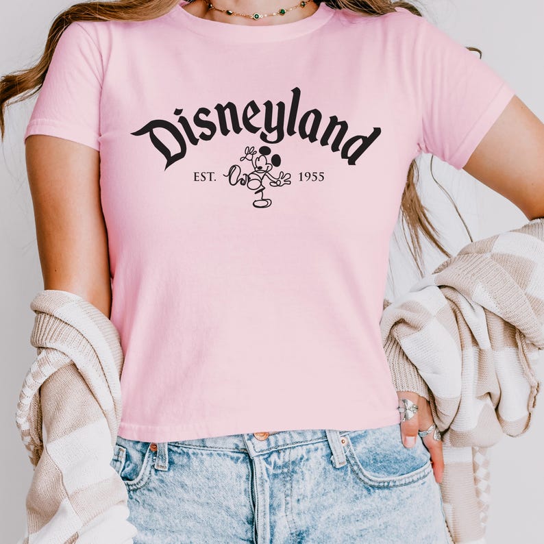 May include: A pale pink t-shirt with the word "Disneyland" in black script. Below the word is a black graphic of Mickey Mouse and the text "EST. 1955". The shirt has short sleeves.