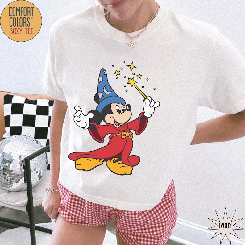 K&ouml;nnte beinhalten: Elfenbeinfarbenes Comfort Colors Boxy Tee mit einem Mickey Mouse-Zauberer-Motiv, das einen Zauberstab mit Sternen h&auml;lt. Das T-Shirt hat einen Rundhalsausschnitt und kurze &Auml;rmel. Dazu werden rot-wei&szlig; karierte Shorts getragen.