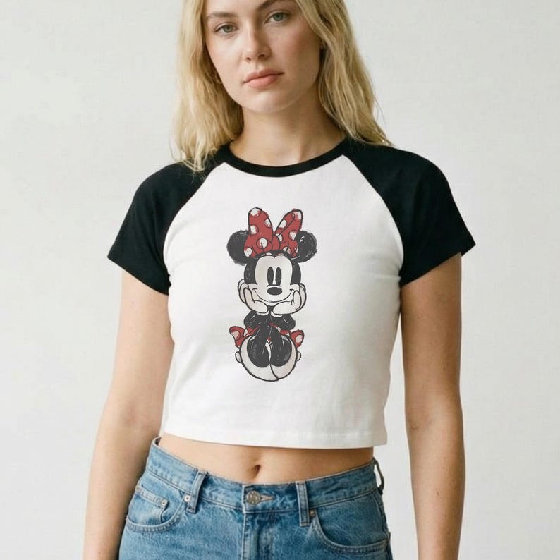 May include: White crop top with black sleeves featuring a Minnie Mouse graphic. The Minnie Mouse illustration has a red bow with white polka dots. The shirt is a baseball style tee.