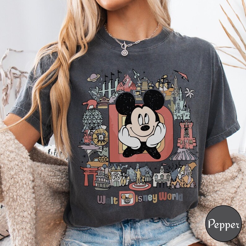 Puede incluir: Camiseta gris oscuro con un gr&aacute;fico colorido de Mickey Mouse y el texto "Walt Disney World". El dise&ntilde;o incluye varios puntos de referencia y atracciones de los parques Disney, con un estilo retro y vintage.