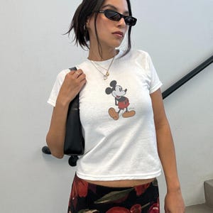 May include: White t-shirt featuring a classic Mickey Mouse graphic. The cartoon character is depicted in black, red, and tan. The shirt is short-sleeved and appears to be made of a soft material.