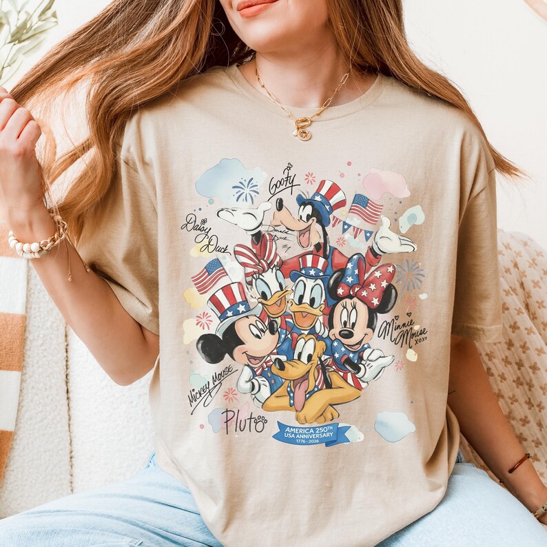 May include: Tan t-shirt featuring a colorful graphic of Disney characters Mickey Mouse, Minnie Mouse, Donald Duck, Daisy Duck, Goofy, and Pluto, celebrating the USA's 250th anniversary. The design includes American flags and fireworks.