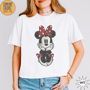 May include: White Comfort Colors boxy tee featuring a distressed graphic of Minnie Mouse. Minnie is depicted with a red polka dot bow, black ears, and a black and white outfit. The shirt has short sleeves and a relaxed fit.