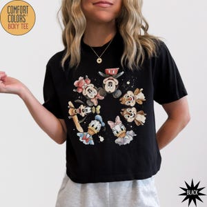 May include: Black Comfort Colors boxy tee featuring a circular graphic of cartoon characters including Mickey Mouse, Minnie Mouse, Donald Duck, Daisy Duck, Goofy, and Chip and Dale. The shirt has short sleeves and a crew neck.