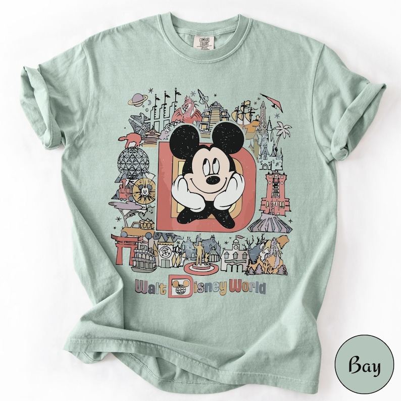 Puede incluir: Camiseta verde claro con un gr&aacute;fico colorido de Mickey Mouse frente a la letra "D" y varios lugares emblem&aacute;ticos de Disney World. Las palabras "Walt Disney World" est&aacute;n impresas debajo. La camiseta tiene las mangas dobladas.