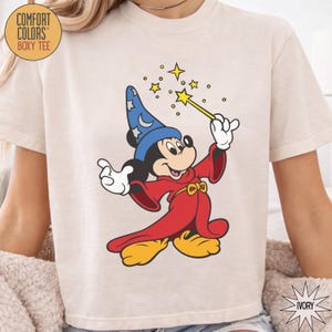 K&ouml;nnte beinhalten: Elfenbeinfarbenes Comfort Colors Boxy-T-Shirt mit einer Grafik von Mickey Mouse als Zauberer. Mickey tr&auml;gt einen blauen Hut mit Sternen, eine rote Robe und gelbe Schuhe. Er h&auml;lt einen Zauberstab mit Sternen. Der Text "COMFORT COLORS" und "BOXY TEE" ist auf dem T-Shirt.