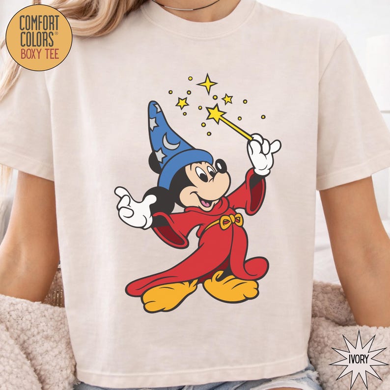 K&ouml;nnte beinhalten: Elfenbeinfarbenes Comfort Colors Boxy-T-Shirt mit einer Grafik von Mickey Mouse als Zauberer. Mickey tr&auml;gt einen blauen Hut mit Sternen, eine rote Robe und gelbe Schuhe. Er h&auml;lt einen Zauberstab mit Sternen. Der Text "COMFORT COLORS" und "BOXY TEE" ist auf dem T-Shirt.
