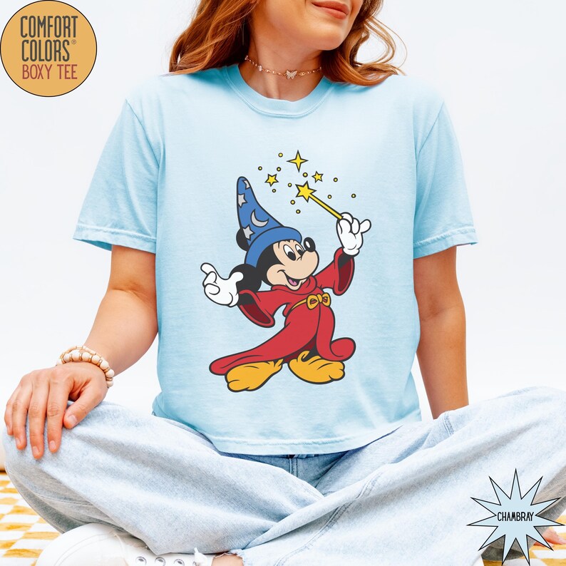 K&ouml;nnte beinhalten: Hellblaues Comfort Colors Boxy-T-Shirt mit einer Grafik von Mickey Mouse als Zauberer, der einen Zauberstab mit Sternen h&auml;lt. Die Zeichentrickfigur tr&auml;gt einen blauen Zaubererhut und eine rote Robe. Das Shirt hat die Farbe Chambray.
