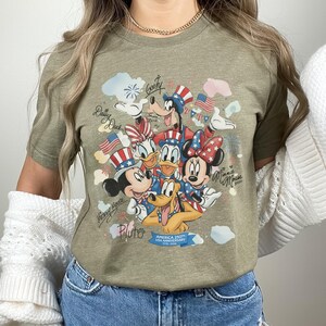 May include: Olive green t-shirt featuring a colorful graphic of Disney characters Mickey Mouse, Minnie Mouse, Donald Duck, Daisy Duck, Goofy, and Pluto. The design includes patriotic elements like American flags and stars, with text that reads "America 250th USA Anniversary 1776-2026."