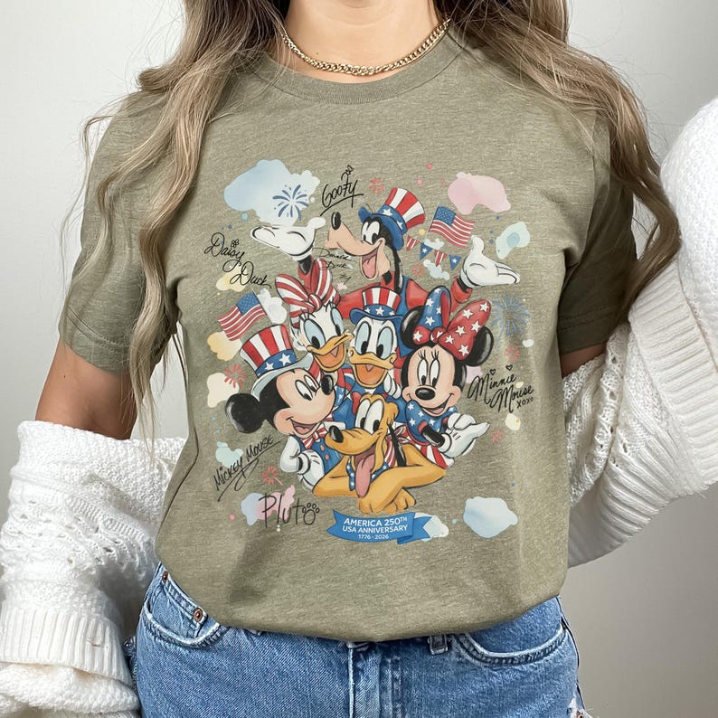 May include: Olive green t-shirt featuring a colorful graphic of Disney characters Mickey Mouse, Minnie Mouse, Donald Duck, Daisy Duck, Goofy, and Pluto. The design includes patriotic elements like American flags and stars, with text that reads "America 250th USA Anniversary 1776-2026."