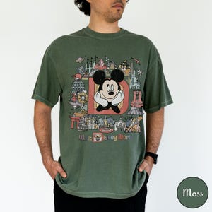 Puede incluir: Camiseta verde musgo con un gr&aacute;fico de Mickey Mouse dentro de una gran letra "D" y el texto "Walt Disney World". El dise&ntilde;o incluye varios iconos del parque en un estilo colorido y vintage. La camiseta es de corte holgado.
