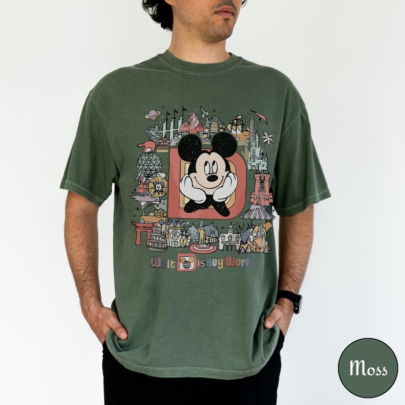 Puede incluir: Camiseta verde musgo con un gr&aacute;fico de Mickey Mouse dentro de una gran letra "D" y el texto "Walt Disney World". El dise&ntilde;o incluye varios iconos del parque en un estilo colorido y vintage. La camiseta es de corte holgado.