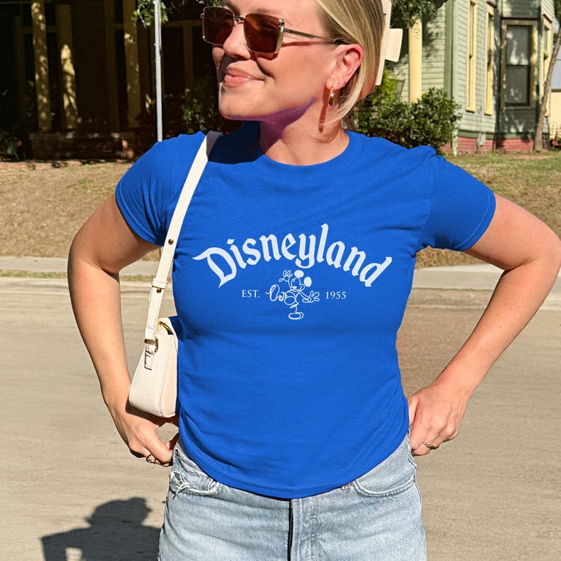 May include: A royal blue t-shirt featuring the word "Disneyland" in white script. Below the word is "EST. 1955" and a Mickey Mouse graphic. The shirt is short-sleeved with a crew neck.