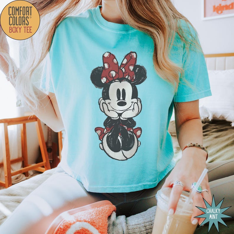 May include: A light blue Comfort Colors boxy tee featuring a distressed graphic of Minnie Mouse. Minnie is depicted with a red polka dot bow and black and white details. The shirt is a relaxed fit.