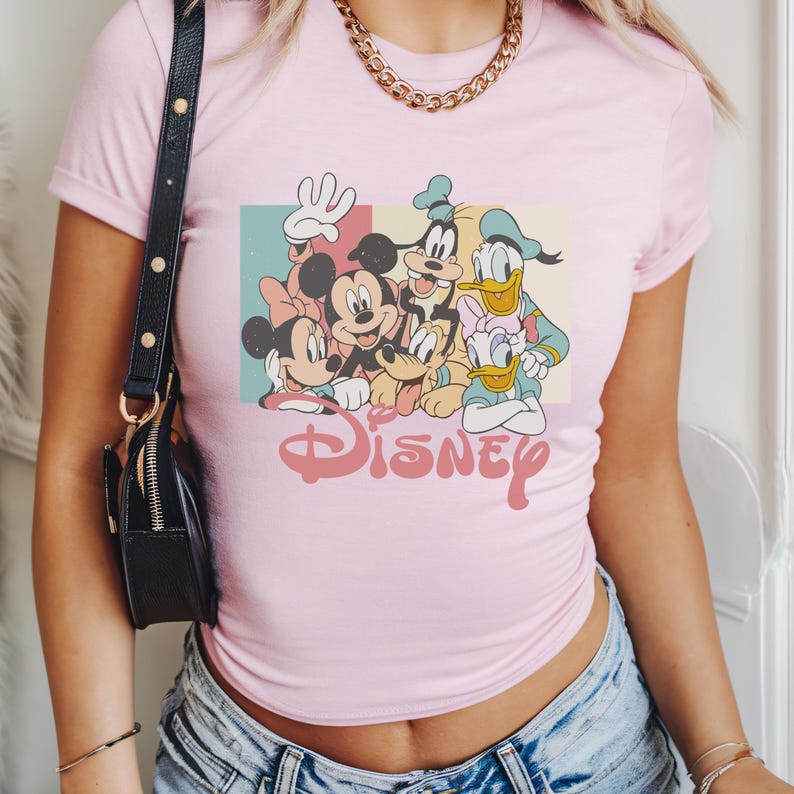 Pu&ograve; includere: T-shirt rosa chiaro con una grafica dei personaggi Disney. Il design include Topolino, Minnie, Pippo, Paperino e Paperina. La parola "Disney" &egrave; stampata in corsivo sotto i personaggi.