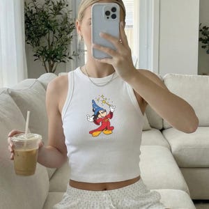 May include: White ribbed tank top featuring a colorful graphic of Mickey Mouse as a wizard, holding a wand with stars. The sleeveless top is a casual style. The image also shows a clear plastic cup with a straw.