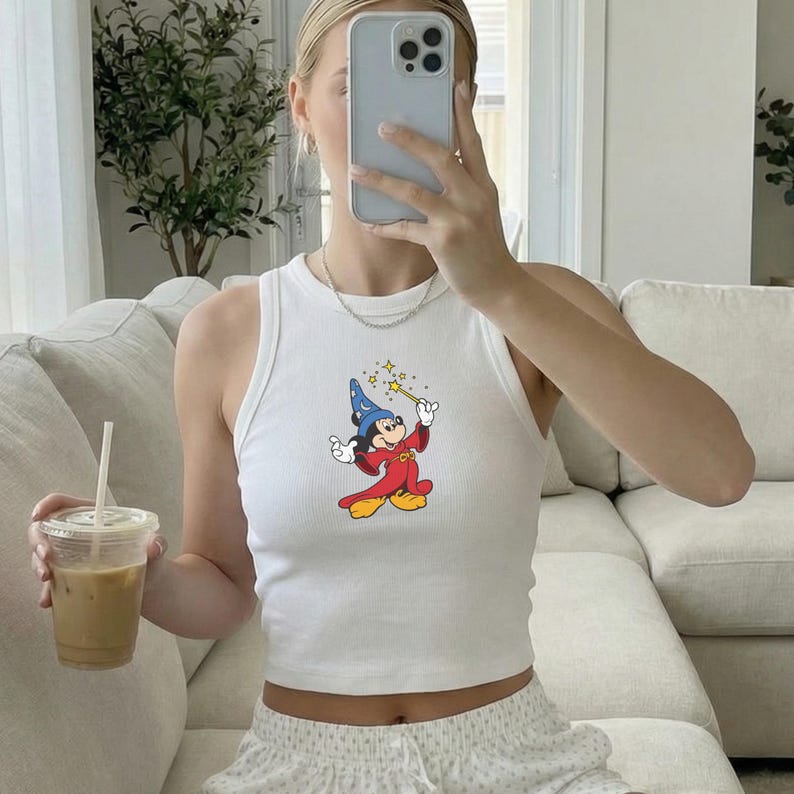 May include: White ribbed tank top featuring a colorful graphic of Mickey Mouse as a wizard, holding a wand with stars. The sleeveless top is a casual style. The image also shows a clear plastic cup with a straw.