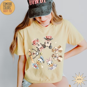 May include: Butter yellow Comfort Colors boxy tee featuring a circular watercolor design of Disney characters including Mickey Mouse, Minnie Mouse, Donald Duck, Daisy Duck, Goofy, and Chip and Dale. The shirt has short sleeves and a relaxed fit.