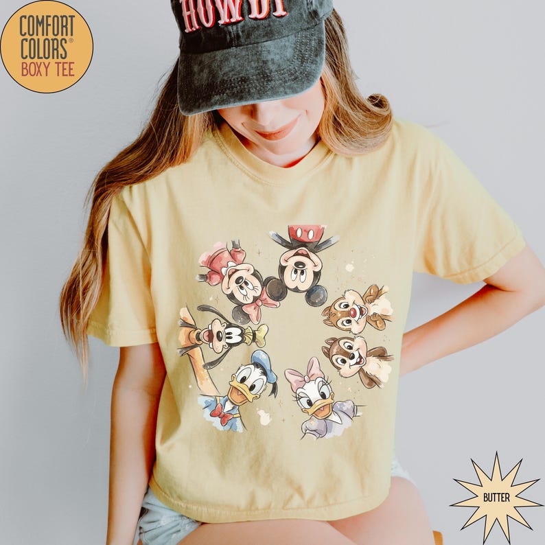 May include: Butter yellow Comfort Colors boxy tee featuring a circular watercolor design of Disney characters including Mickey Mouse, Minnie Mouse, Donald Duck, Daisy Duck, Goofy, and Chip and Dale. The shirt has short sleeves and a relaxed fit.