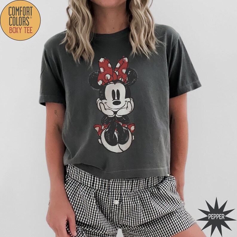 May include: Dark gray Comfort Colors boxy tee featuring a distressed Minnie Mouse graphic with a red bow and polka dots. The shirt has short sleeves and is paired with black and white checkered shorts. The shirt has the text "Comfort Colors Boxy Tee" on the upper left.