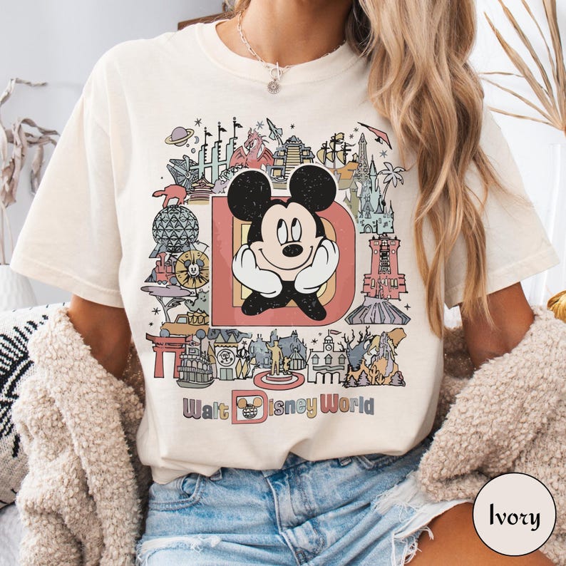 Puede incluir: Camiseta de color marfil con un gr&aacute;fico colorido de Mickey Mouse dentro de una gran letra "D" y varios monumentos de Disney World. Las palabras "Walt Disney World" est&aacute;n impresas debajo. La camiseta tiene un corte relajado.