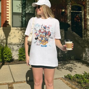 May include: White t-shirt featuring a colorful graphic of Disney characters, including Mickey Mouse, Minnie Mouse, Donald Duck, and Pluto, with patriotic elements like flags and fireworks. The shirt has a crew neck and short sleeves.