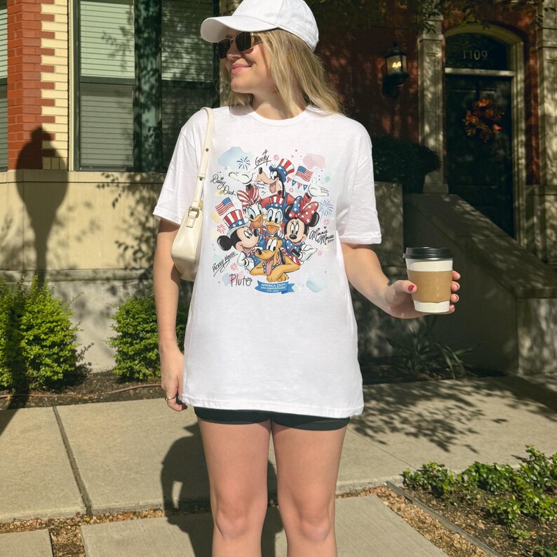 May include: White t-shirt featuring a colorful graphic of Disney characters, including Mickey Mouse, Minnie Mouse, Donald Duck, and Pluto, with patriotic elements like flags and fireworks. The shirt has a crew neck and short sleeves.