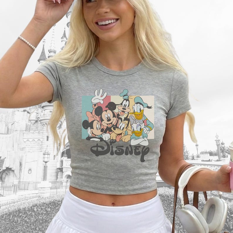 Pu&ograve; includere: Un crop top grigio con una grafica colorata dei personaggi Disney, tra cui Topolino, Minnie, Pippo, Paperino e Pluto. La parola "DISNEY" &egrave; stampata in un font vintage sotto i personaggi. La maglietta ha maniche corte.