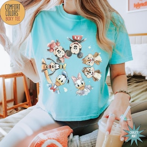 May include: A light blue Comfort Colors boxy tee featuring a circular design of Disney characters, including Mickey Mouse, Minnie Mouse, Donald Duck, Daisy Duck, Goofy, and Chip and Dale. The shirt has a relaxed fit and a crew neck.