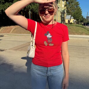 May include: A red t-shirt featuring a graphic of Mickey Mouse. The shirt has short sleeves and is paired with light blue jeans. A small, white purse with a shoulder strap is visible.