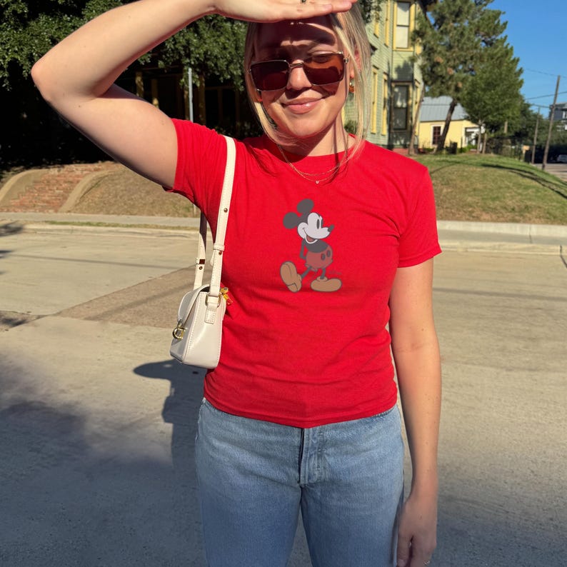 May include: A red t-shirt featuring a graphic of Mickey Mouse. The shirt has short sleeves and is paired with light blue jeans. A small, white purse with a shoulder strap is visible.