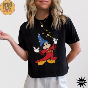 K&ouml;nnte beinhalten: Schwarzes Comfort Colors Boxy-T-Shirt mit einer Grafik von Mickey Mouse als Zauberer, der einen blauen Hut und eine rote Robe tr&auml;gt und einen Zauberstab mit Sternen h&auml;lt. Das Shirt hat eine schwarze Sternengrafik unten rechts.