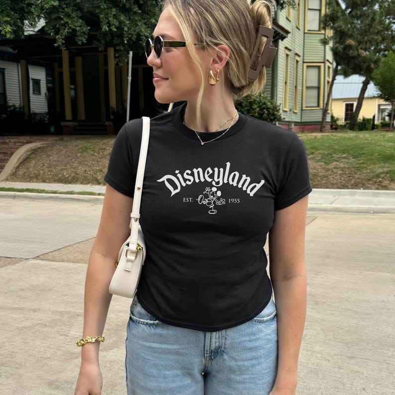 May include: Black crew neck t-shirt with the word "Disneyland" in white script. The shirt features a Mickey Mouse graphic and the text "EST. 1955". The person is wearing light blue jeans and sunglasses.