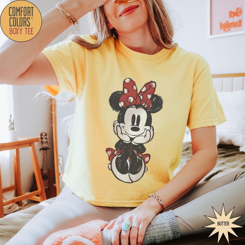 May include: Yellow Comfort Colors boxy tee featuring a distressed Minnie Mouse graphic with a red polka dot bow. The shirt has short sleeves and a relaxed fit. The text "COMFORT COLORS BOXY TEE" is in a circle on the top left.