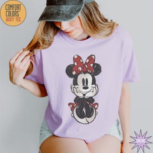 May include: A light purple Comfort Colors boxy tee featuring a distressed Minnie Mouse graphic. Minnie is depicted in black, red, and white, with a red polka dot bow. The shirt has short sleeves and a relaxed fit. The text "COMFORT COLORS" and "BOXY TEE" are printed on the upper left.
